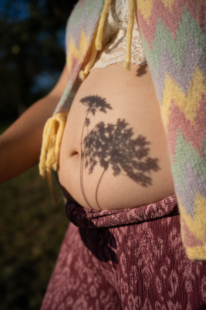 Detailaufnahme Babybauch mit Schattenspiel einer Blume, buntes Oberteil im Sonnenlicht