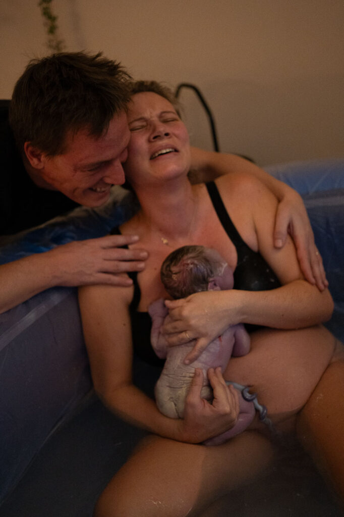 Frau hält Neugeborenes im Geburtspool, Mann umarmt sie überglücklich, emotionale Geburtsfotografie Esslingen