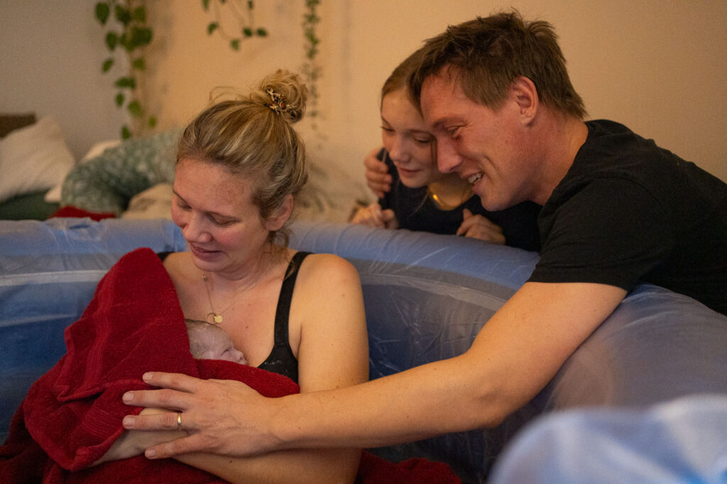 Frau sitzt in Geburtspool mit Neugeborenem im Handtuch auf ihrer Brust, Mann und älteres Kind betrachten das Baby, emotionale Geburtsfotografie Esslingen