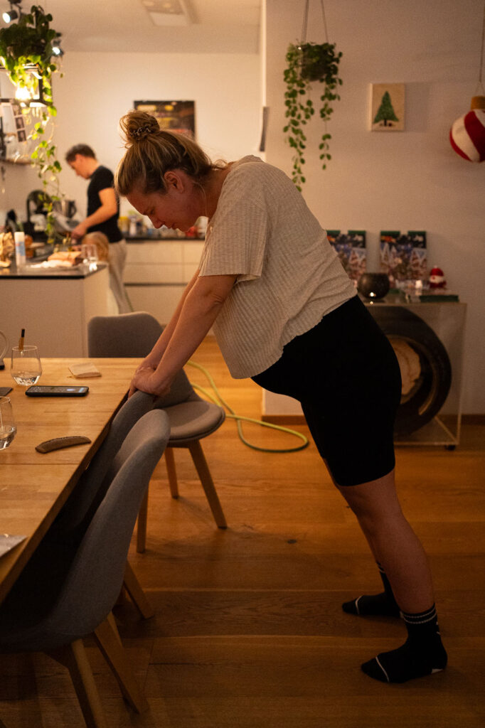 Frau stützt sich auf Stuhl und veratmet Wehe, Mann im Hintergrund, Geburtsfotografie Hausgeburt Esslingen