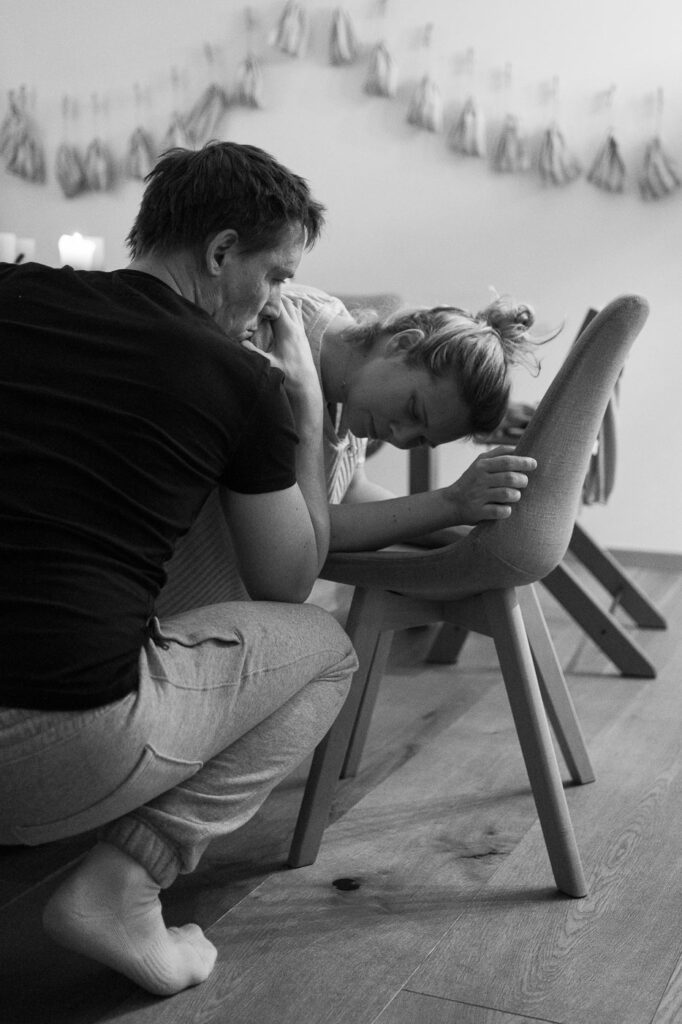 Frau stützt sich kniend auf Stuhl, Mann stützt sie, Geburtsfotografie Hausgeburt Esslingen