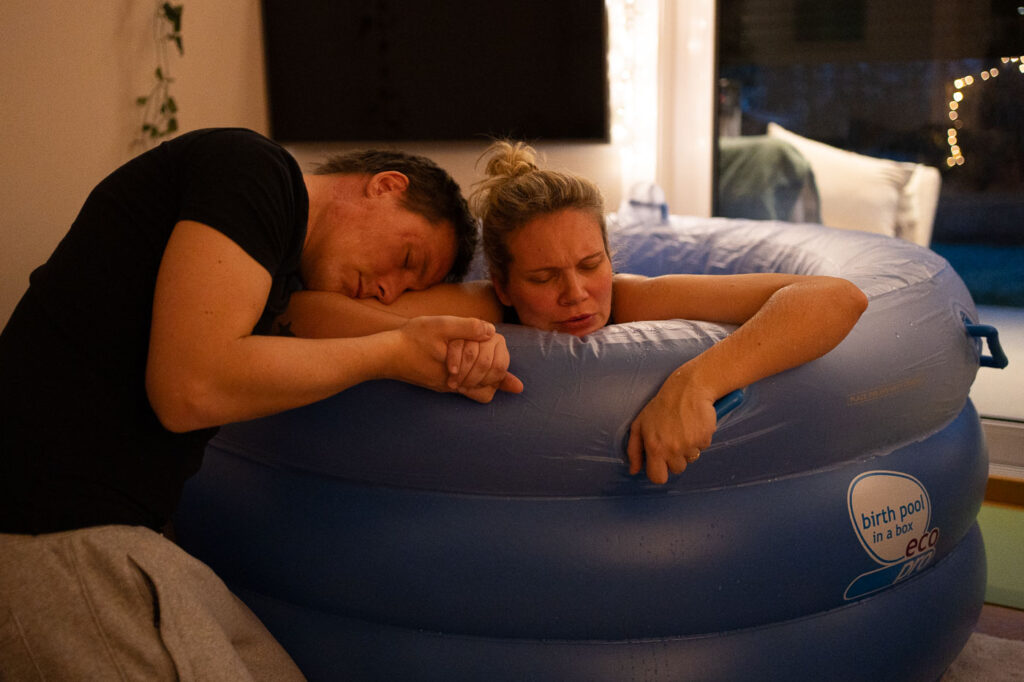 Frau stützt sich auf Geburtspool-Rand auf und veratmet Wehe, Mann hält sie kniend fest, Geburtsfotografie Stuttgart, Geburtsfotografie Esslingen
